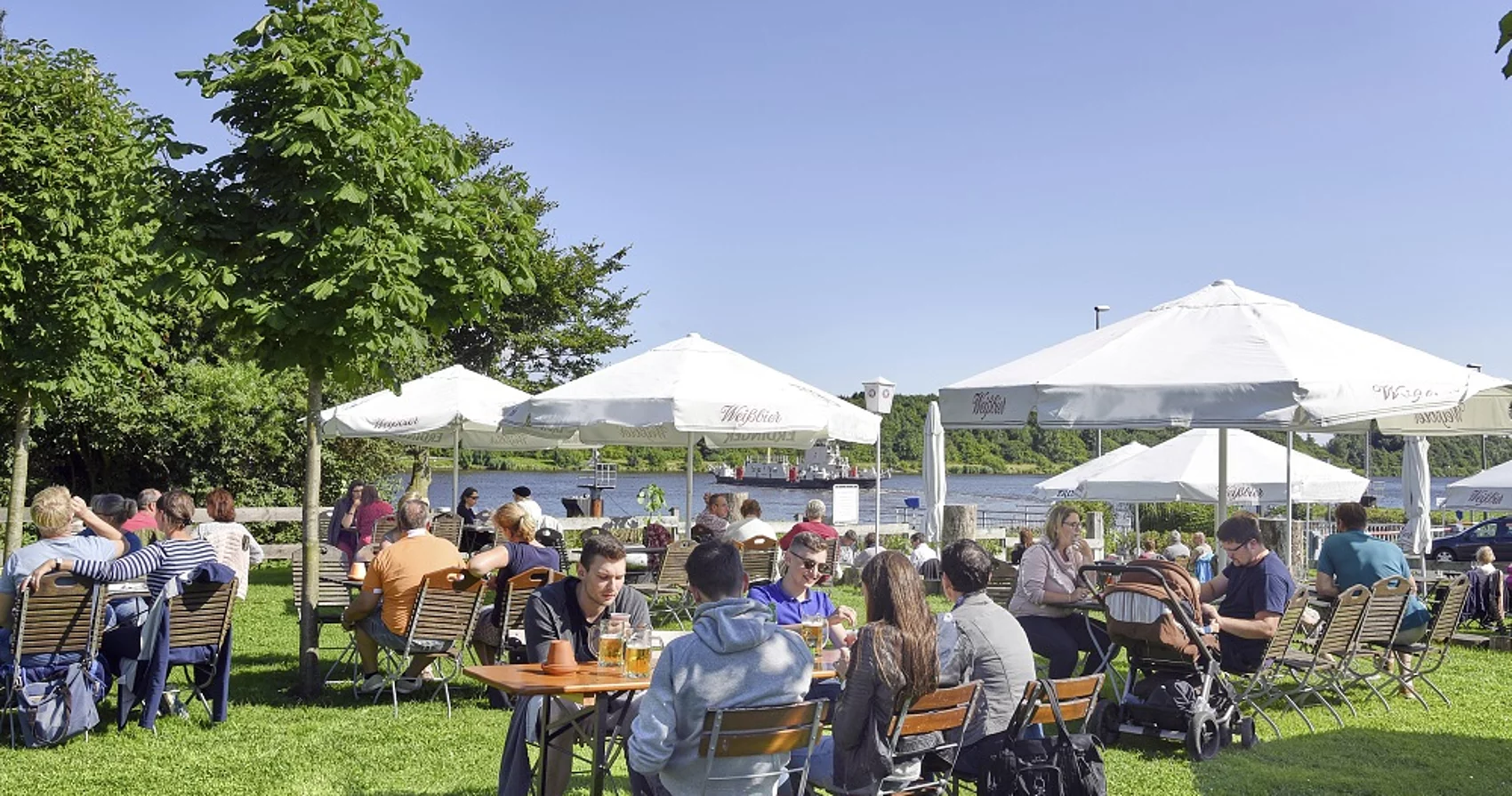 Auf einer grünen Wiese sitzen Menschen gemütlich vor einem Restaurant und genießen ein Getränk mit Blick auf den Kanal.