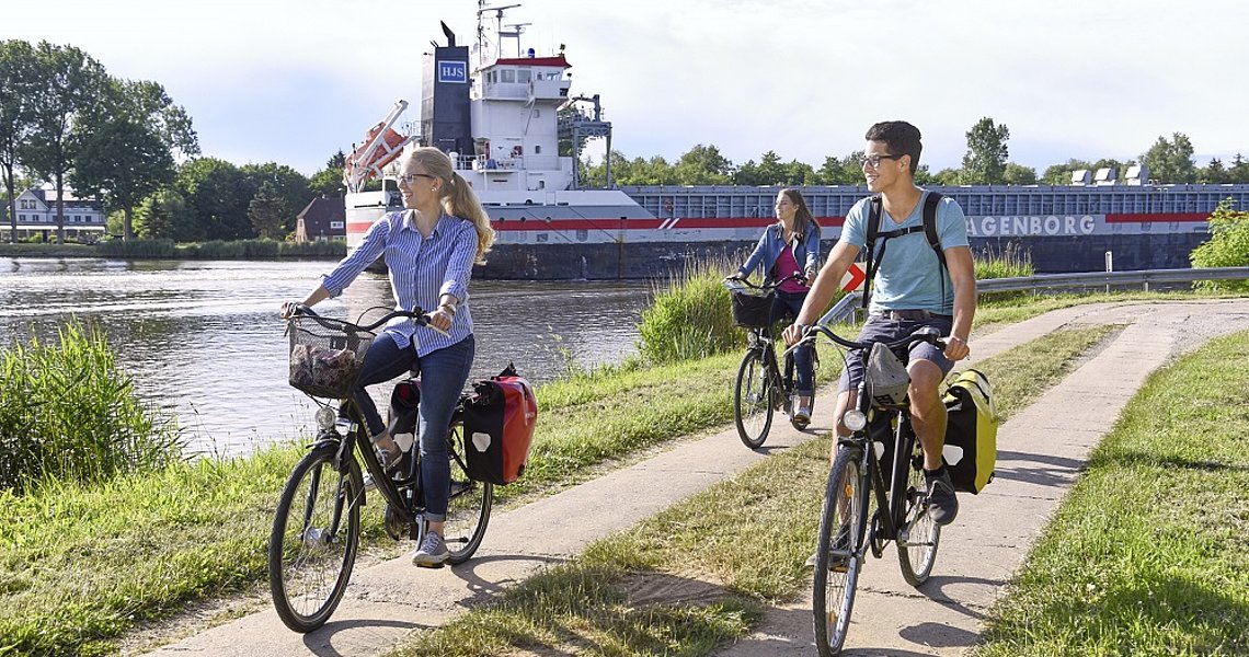 Fahrradfahrer fahren am Kanal entlang. Im Hintergrund fährt ein Schiff auf dem Kanal.