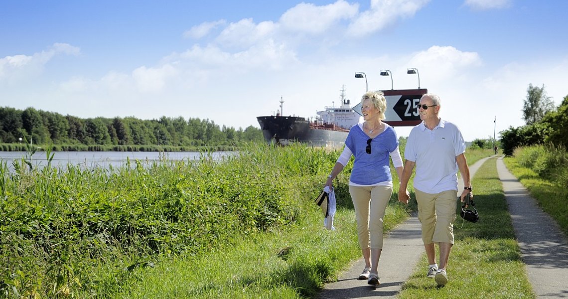 Zwei Menschen spazieren auf dem Weg neben dem Kanal. Im Hintergrund ist ein Schiff zu sehen.