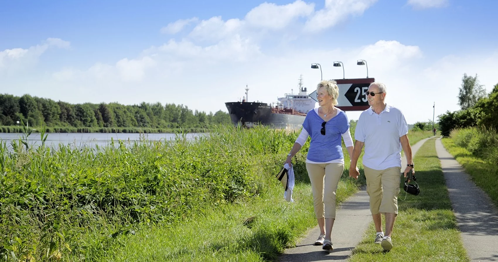 Zwei Menschen spazieren auf dem Weg neben dem Kanal. Im Hintergrund ist ein Schiff zu sehen.