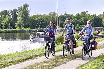 Drei Fahrradfahrer radeln auf dem Plattenweg parallel zum Nord-Ost-See-Kanal, auf dem ein Segelschiff zu sehen ist.