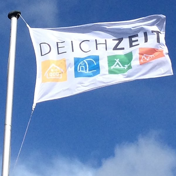 eine weiße Flagge mit dem Schriftzug DEICHZEIT flattert vor blauem Himmel