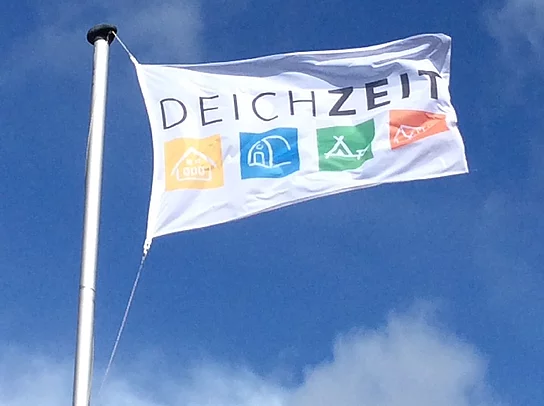 eine weiße Flagge mit dem Schriftzug DEICHZEIT flattert vor blauem Himmel